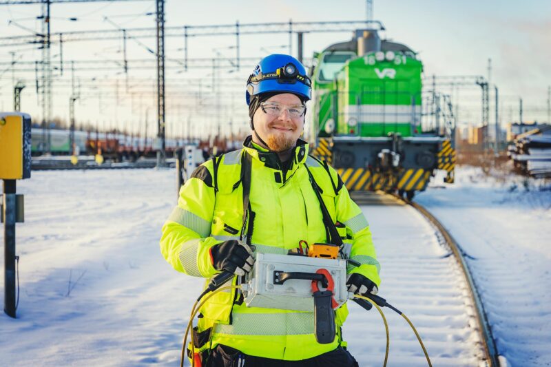 Installatören av säkerhetsanordningar Jonne Lähde står iklädd hjälm och arbetsrock och tittar leende in i kameran. I bakgrunden syns ett VR-lok.